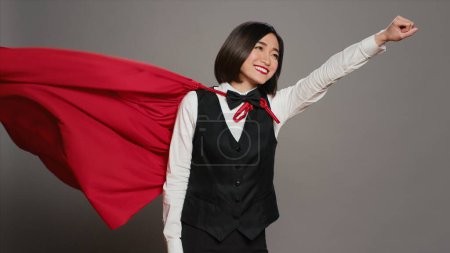 Photo for Front desk operator feeling strong with a superhero cape on camera, posing with confidence and showing power in studio. Hotel concierge wearing a red hero cloak, hospitality industry. Camera A. - Royalty Free Image