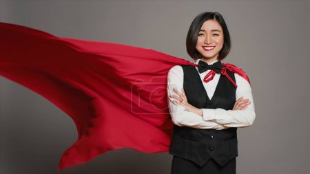 Téléchargez les photos : Administrateur de l'hôtel portant un manteau de héros rouge à la caméra, se sentant fort et puissant pour aider les clients. réceptionniste asiatique posant avec confiance à l'aide d'une cape de super-héros sur fond gris. Caméra A. - en image libre de droit