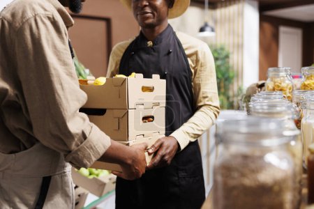 Landwirt liefert Bio-Gemüse an Verkäufer, die an Kunden verkaufen, die sich auf nachhaltiges Leben in einem Laden ohne Abfall konzentrieren. Ein Mann reicht Kisten mit frisch gepflückten Naturprodukten an einen Ladenbesitzer weiter.