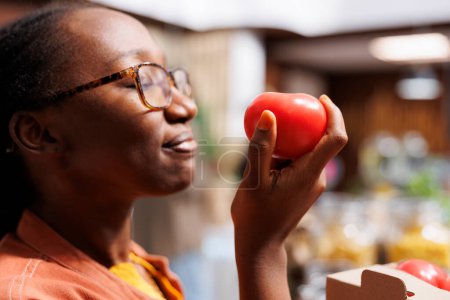 Selective focus on an African American woman at eco friendly market, admiring fresh, organic produce. Health-conscious, female customer inhales natural scents, supporting local, sustainable practices.