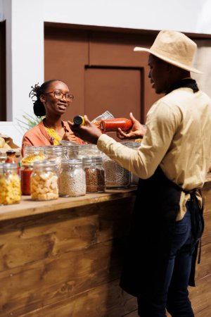 Foto de Un comerciante afroamericano ecológico vende comestibles frescos y orgánicos. Una joven clienta indaga sobre productos nutritivos de origen local en la tienda libre de plástico. - Imagen libre de derechos