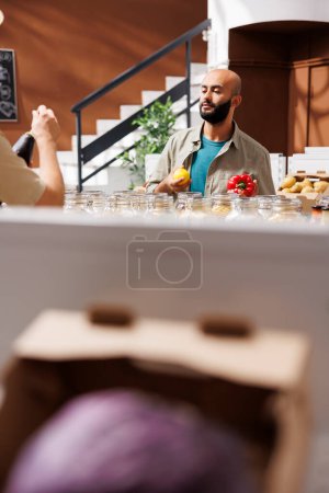 Photo for Middle Eastern male customer holds a lemon in one hand and red pepper in another, admiring them both. Young bearded client browsing in an environmentally friendly grocery store. - Royalty Free Image