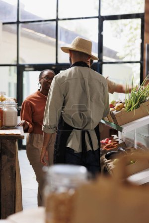 Foto de Shopkeeper proporciona asesoramiento experto a diversas parejas mientras compran alimentos frescos, saludables y naturales. Store enfatiza las opciones ecológicas, promoviendo pequeñas empresas que valoran la sostenibilidad. - Imagen libre de derechos