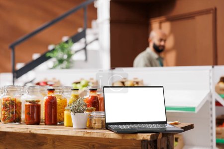 Wireless computer displaying white screen in bio shop filled with freshly harvested fruits and vegetables. Digital laptop showing isolated chromakey mockup template in zero waste store.