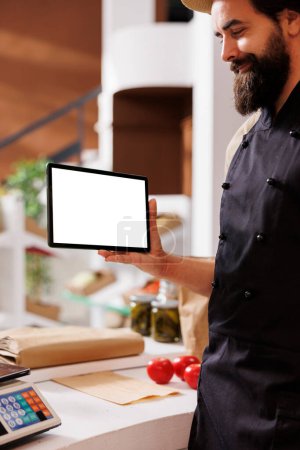 While holding tablet with isolated white screen, male shopkeeper waits to assists customers. Caucasian male seller gripping a digital device horizontally with blank copyspace mockup template.