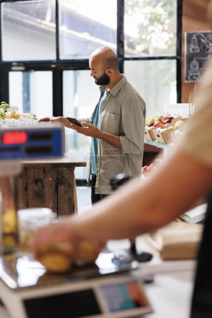 Ein arabischer männlicher Kunde surft im Internet, während er sich einen bestimmten Abschnitt in einem umweltfreundlichen Lebensmittelgeschäft ansieht. Moderne Technologie im nachhaltigen Supermarkt.