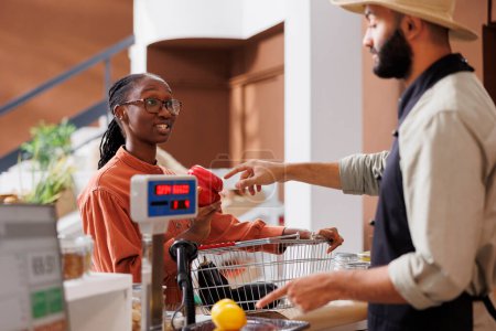 Mujer negra en el mostrador de pago con una cesta de la compra llena de productos alimenticios cultivados localmente para que el vendedor pese. Cajero masculino ayudando a cliente afroamericano con sus comestibles.