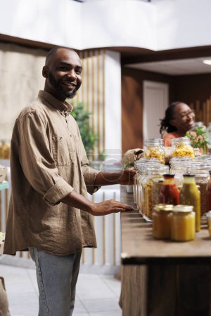Photo for In a modern grocery shop, a happy African American man looks at the camera. The shelves feature reusable packaging, eco friendly products, and a selection of items. - Royalty Free Image