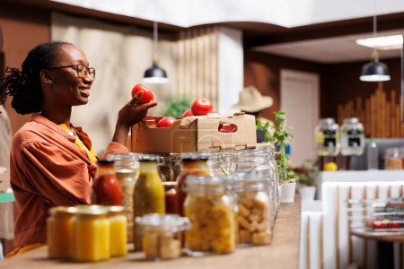 Foto de La foto se centra en la sonriente mujer negra en una tienda de bioalimentos que examina un tomate orgánico cultivado en la granja. Cliente de etnia afroamericana admira alegremente productos frescos en el mercado ecológico. - Imagen libre de derechos