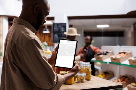 Chico afroamericano hojea los artículos de bio-alimentos, frascos y alimentos en los estantes mientras lleva una tableta del teléfono con una plantilla de cromakey. Hombre negro agarrando un dispositivo digital con una pantalla blanca.