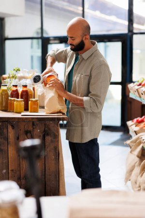 Foto de Hombre de Oriente Medio llenando una bolsa de papel marrón con lentejas naranjas. Joven vertiendo granos en la bolsa, comprando productos saludables de contenedores reutilizables con productos orgánicos a granel. - Imagen libre de derechos