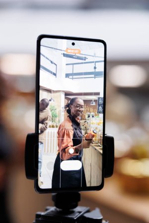 Foto de El dueño de la tienda afroamericana utiliza el teléfono celular y el selfie stick para vlogging, promoviendo artículos sostenibles, saludables y naturales. Alimentos orgánicos y frescos en frascos de vidrio, atractivos para los clientes eco-conscientes. - Imagen libre de derechos