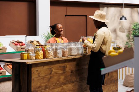 Propietario de tienda masculina discutiendo frutas y verduras frescas en el mercado local con una mujer negra sonriente. Vendedor ofrece alimentos básicos despensa orgánica y productos a granel libres de plástico, la promoción de productos saludables.