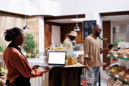 El dueño de una tienda de comestibles afroamericana mira el portátil con una pantalla blanca en blanco. Mujer joven examinando bienes durante el funcionamiento de un PC inalámbrico con cromakey plantilla copyspace. Productos locales sostenibles.