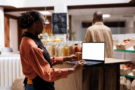 In a grocery store, African American female vendor examines the products with laptop displaying blank chromakey template. The eco friendly shop shows a variety of reusable containers and glass jars.