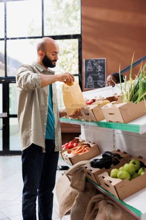 Middle eastern man explores modern eco friendly grocery store, browsing a variety of fresh, organic products with sustainable packaging. Male customer putting fresh potatoes in brown paper bag.