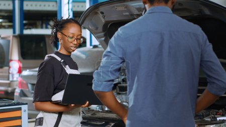 Photo for African american mechanic in repair shop using laptop to calculate repairments invoice for client after fixing car malfunctions. Woman using device to determine final costs after servicing vehicle - Royalty Free Image