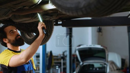 Photo for Precise mechanic underneath vehicle on overhead lift in garage, checking components. Employee using work light to make sure automotive underbody is in perfect condition, doing close examination - Royalty Free Image