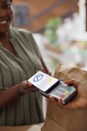 Photo for An African American woman smiles as she pays for freshly harvested organic produce using her mobile phone at a local marketplace. Convenient and eco-friendly shopping with contactless technology. - Royalty Free Image