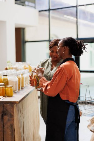 African american vendor showcasing her products in zero waste supermarket, selling organic bio food to interested buyer. Storeowner showing her fresh bulk products merchandise during event.