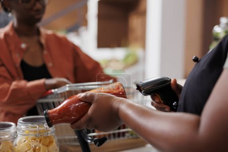 Foto detallada mostrando un cajero afroamericano escaneando una botella de salsa de pasta casera para los clientes. Una vista cercana de una vendedora que utiliza un escáner de código de barras en el mostrador de pago.