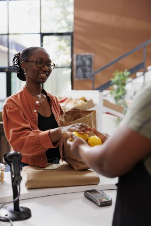 African American women presenting recently picked lemons at the cashier desk. Image showcasing a female customer offering fruits to a black vendor to weigh.