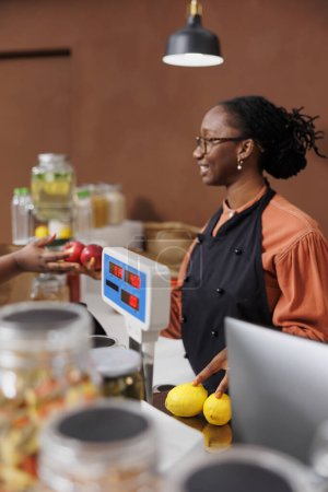 Mujer negra ayuda a un cliente en una tienda de comestibles, la promoción de productos cultivados localmente y orgánicos. Comunican y utilizan la tecnología para un proceso de pago eficiente en el mercado ecológico.
