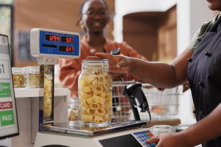 Black woman with spectacles waiting while vendor uses digital scale to measure glass container filled with fresh pasta. African american seller weighing jar of freshly made spaghetti for customer.