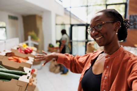 Foto de Vlogger hembra negro mostrando varios productos cultivados localmente de tienda ecológica. Mujer afroamericana en videollamada apuntando hacia frutas y verduras recién cosechadas en la tienda de comestibles. - Imagen libre de derechos