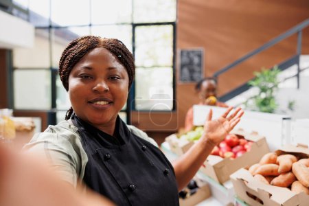 Mujer negra vlogging sobre la vida saludable en la tienda de comestibles local, mostrando productos de alimentos biológicos. Shopkeeper usa smartphone para compartir su mensaje como creadora de contenido y bloguera en una tienda ecológica.