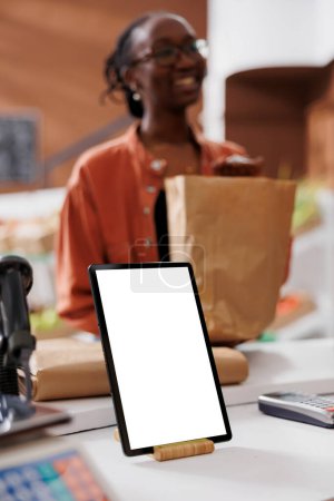 Closeup of digital tablet with blank white screen vertically placed on checkout counter at sustainable bio food market. Device displaying copy space mockup template while customer carries grocery bag.