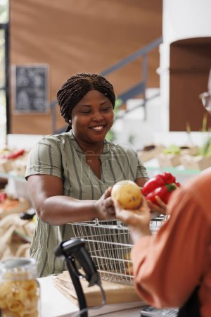 Téléchargez les photos : Heureuse femme afro-américaine faisant des achats individuels pour des légumes biologiques cultivés localement pour des recettes saines. Jeune consommateur donnant au vendeur local les produits frais pour le pesage et l'emballage. - en image libre de droit