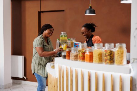 Afroamerikanische Kundin diskutiert mit einer Verkäuferin an der Kasse über umweltfreundliches Einkaufen, während sie Gläser mit Müsli und Honig zum Scannen überreicht. Schwarze Frau kauft frische Bio-Lebensmittel.