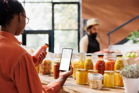 African american customer analyzes glass bottle filled with red sauce while also holding mobile device with white screen in grocery store. Black woman grasping cellphone with blank mockup template.
