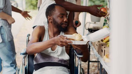 Kostenlose Mahlzeiten und Obst werden einem an den Rollstuhl gefesselten Afrikaner in Not gegeben. Behinderte, arme und obdachlose Menschen werden von einer gemeinnützigen Ernährungsorganisation versorgt. Nahaufnahme, Stativaufnahme.