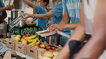 Bei der Essensausgabe erhalten arme Rollstuhlfahrerinnen kostenlose Lebensmittel und Proviant. Multiethnische Freiwillige in blauem T-Shirt verteilen frisches Obst und warme Mahlzeiten an weniger Privilegierte. Nahaufnahme, Stativ.
