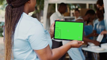 Photo for Young black woman clutching digital tablet with isolated mockup template for charitable messaging. African american volunteer carrying smart gadget with blank green screen. Over the shoulder tripod. - Royalty Free Image