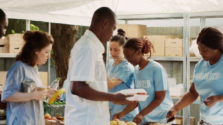 Téléchargez les photos : Les femmes bénévoles multiraciales servent des repas et des produits non périssables aux personnes affamées et sans abri. Des œuvres de charité amicales distribuent gratuitement de la nourriture aux pauvres et aux nécessiteux. Trépied. - en image libre de droit