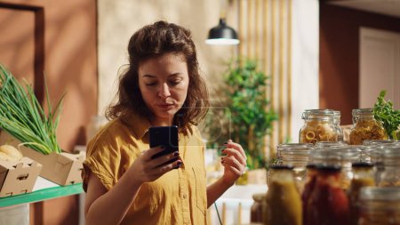 Vegan woman checking zero waste supermarket reviews on smartphone before doing shopping. Customer using cellphone in environmentally friendly local neighborhood grocery shop