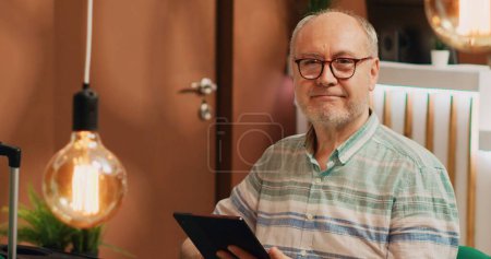 Older man tourist on lounge area sofa, surfing the web on digital tablet. Person holding device to browse internet waiting to see room at five star hotel on vacation, checks online reservation.