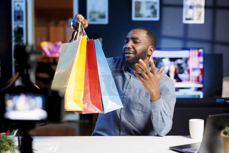 Photo for Blogger doing shopping haul, holding colorful bags in hand, showing subscribers purchases he recently got. Internet star presenting fanbase new acquisitions, filming himself in apartment - Royalty Free Image