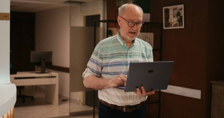 Older person searching for places to visit on laptop, using wireless connection at hotel before registering procedure. Retired man travelling abroad on vacation, browsing online information.