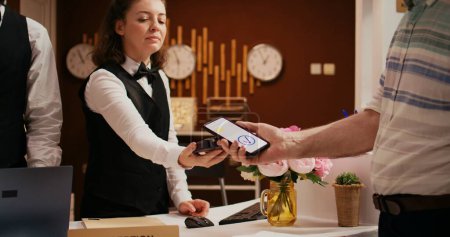 Retired guest using mobile payment with nfc technology at pos terminal, paying for hotel room at reception front desk. Old man registering at holiday retreat, making electronic transaction.