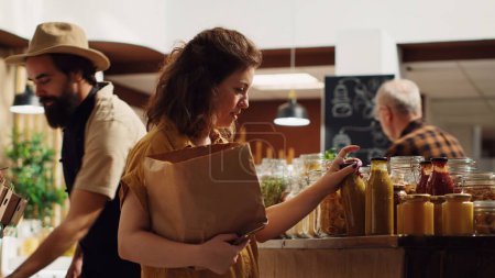 Photo for Woman adding additives free natural pantry essentials and vegetables in paper bag, getting product recommendations from vendor in zero waste supermarket. Retail clerk helping client in grocery shop - Royalty Free Image