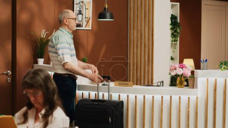 Elderly guest arrives in hotel lobby, greeted by skilled staff members at reception. Senior person carrying luggage for international vacation, preparing for check in, ring service bell.