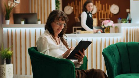 Hotel guest using modern device in lounge area, waiting for check in and navigating on online webpage. Elderly woman browsing internet after arriving at five star holiday resort. Handheld shot.