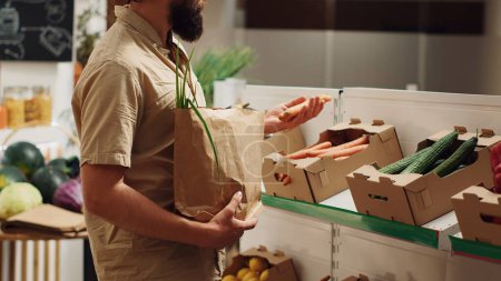 Vegan man in zero waste supermarket using nonpolluting paper bag while shopping for natural vegetables. Customer in low carbon footprint local grocery shop with no single use plastics policy