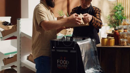 Lieferant, der den Kunden Bio-Lebensmittel ohne Abfall zur Verfügung stellt, unterstützt von einer älteren Einzelhandelskauffrau beim Befüllen des Thermorucksacks. Mann bringt umweltfreundliche Lebensmittel zu den Kunden