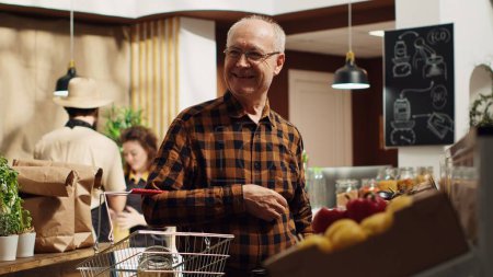 Porträt eines lächelnden alten Kunden in einem Supermarkt, der mit seinem Einkaufskorb Massengüter in wiederverwendbaren Glascontainern kauft. Älterer Mann im örtlichen Lebensmittelgeschäft kauft Grundnahrungsmittel