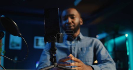 Photo for Close up of cellphone on holder and microphone used by online star in dimly lit home studio to film video for online platforms. Phone being used by african american influencer to record footage - Royalty Free Image